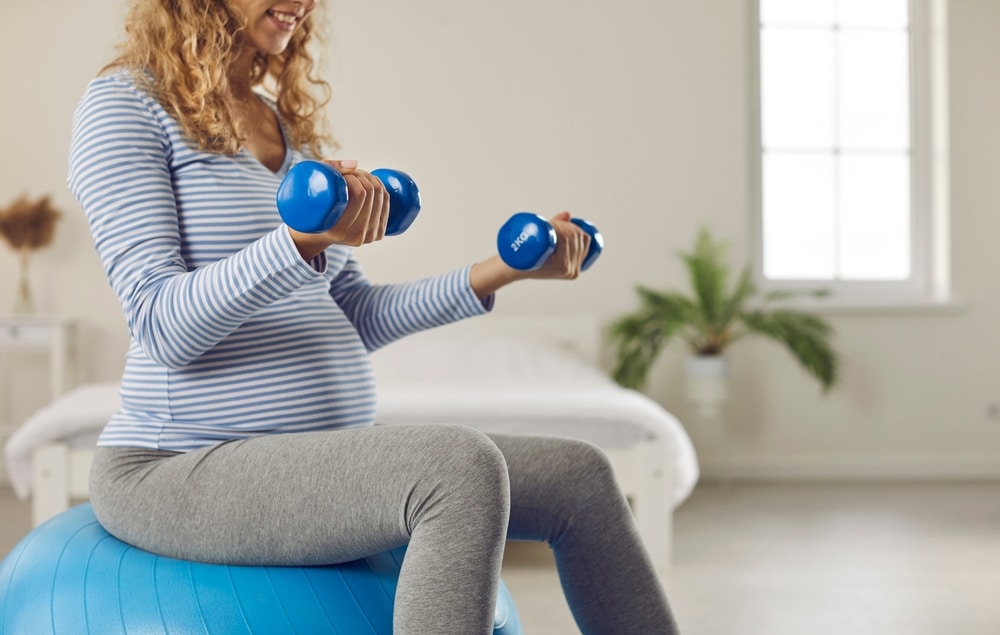 Pregnant woman sitting on fit ball and doing physio therapy exercises with light weights. Active lady balancing on fitball with dumbbells in hands, cropped shot.