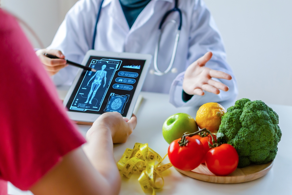 Nutritionist female doctor using digital mobile tablet with virtual graphic icon diagram and vegetable and fruit with patient on desk at office hospital, nutrition, food science, healthy food concept