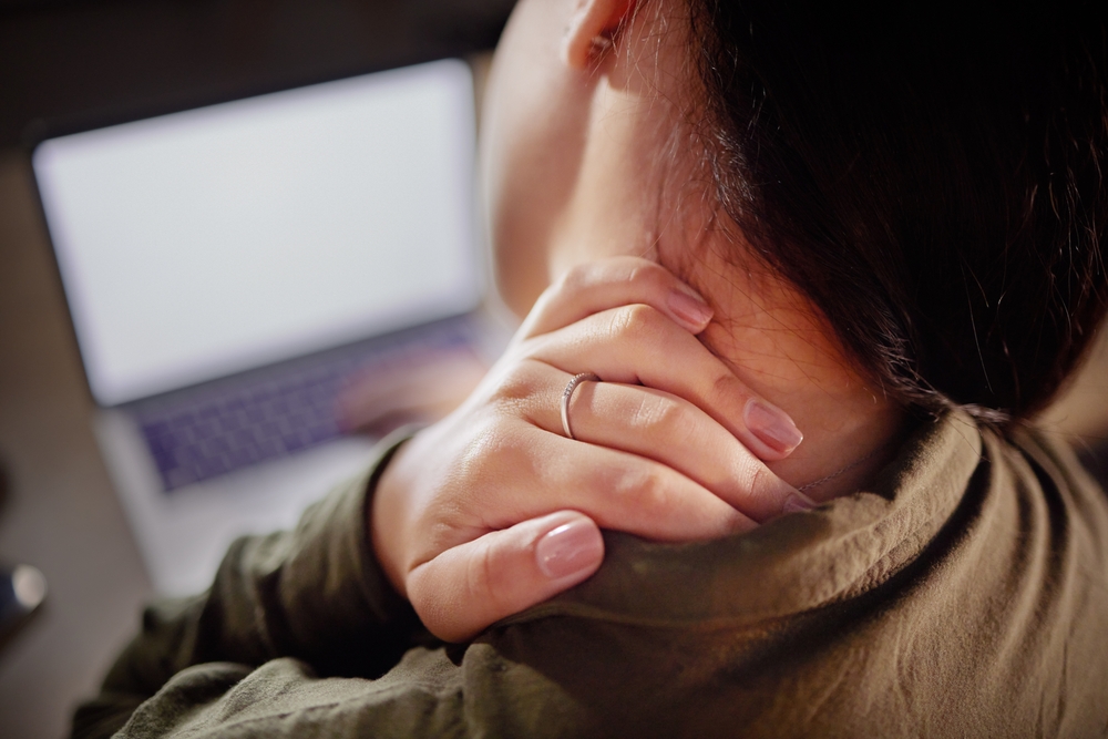 A woman grimaces in discomfort, holding her neck while working on a laptop, indicating neck pain from prolonged use.